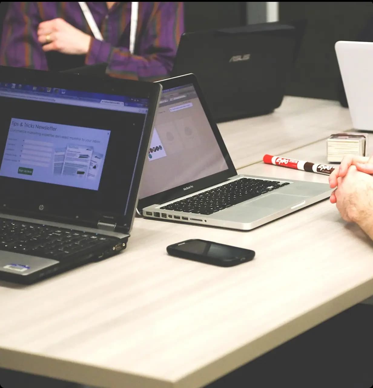 Business meeting at a conference table with open laptops, a smartphone, and a marker; people discussing in the background.