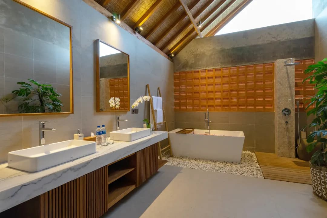 Ensuite bathroom of bedroom three at Jiwoku Villas Canggu Bali featuring double sinks, freestanding bathtub with flower petals, tropical plants, and natural skylight under bamboo ceiling
