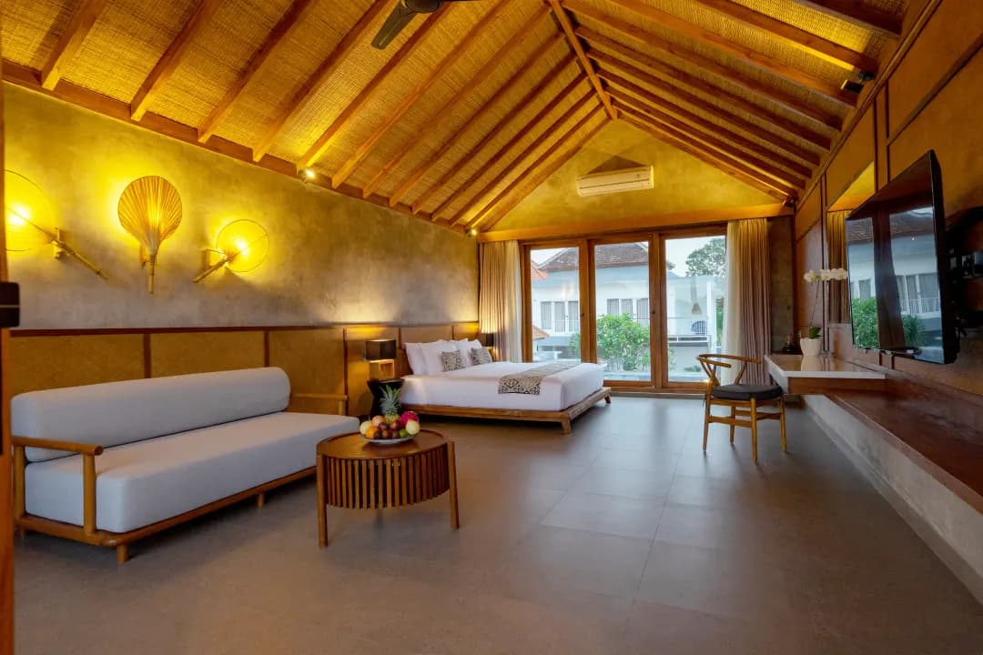 Bedroom three at Jiwoku Villas Canggu Bali featuring wooden bed, lounge sofa, bamboo ceiling, soft warm lighting, and large glass doors overlooking tropical courtyard