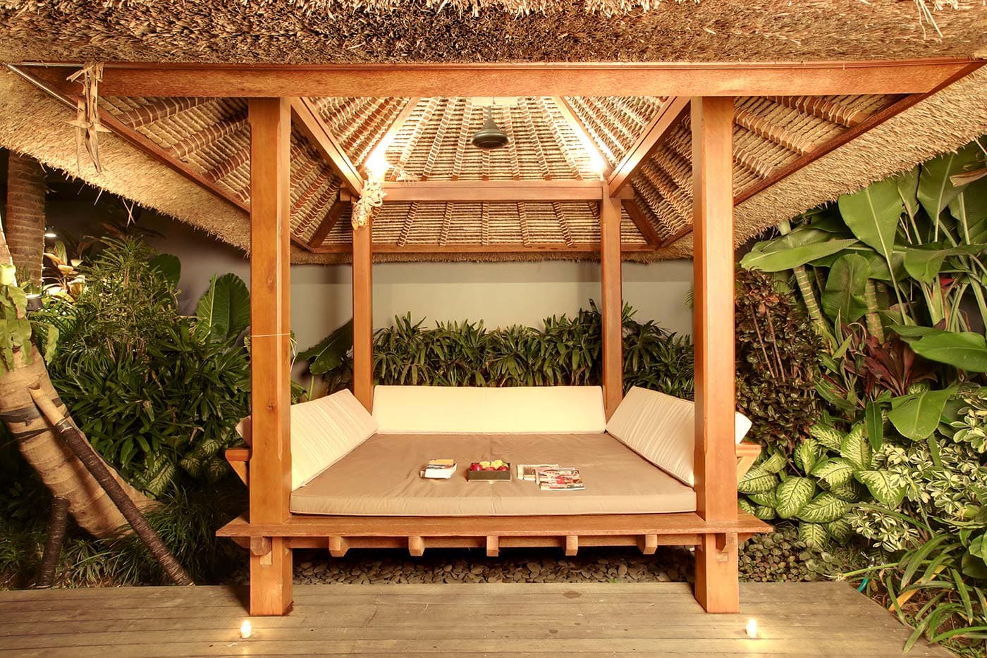 Tropical-style wooden pavilion with a thatched roof and a large central daybed, surrounded by lush green plants, creating a relaxing and natural atmosphere.