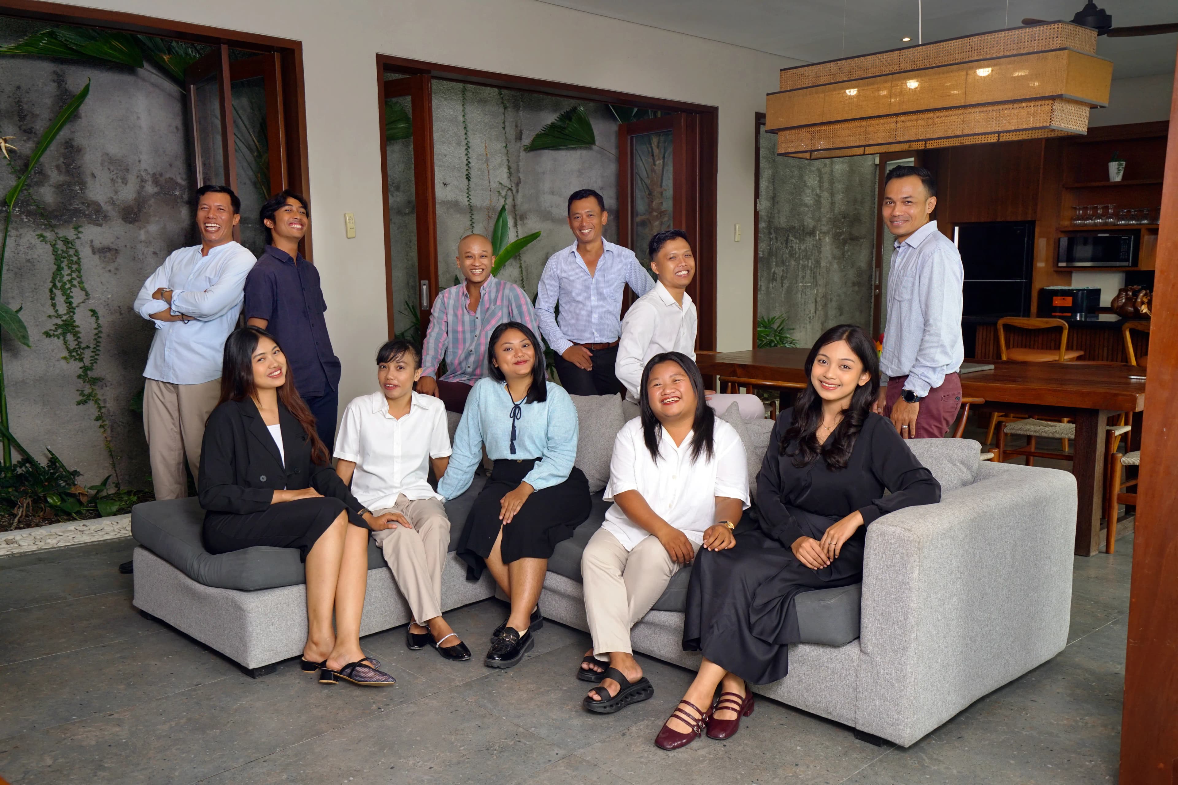 Eclectic Villas team group photo — 11 staff smiling in a modern Bali villa living room, seated and standing around a grey sofa